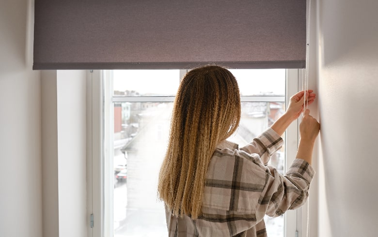 a woman stands in front of a window pulling down a blackout roller blind