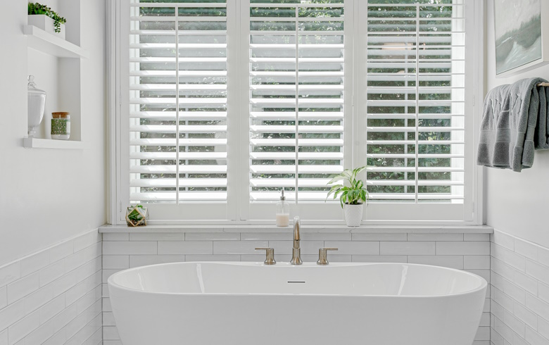 White PVC shutters in a white tiled bathroom with a free-standing bath.